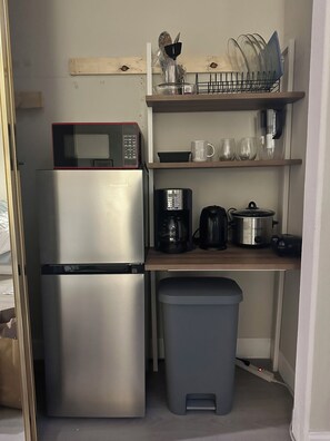 Eine kleine Küchenzeile mit Edelstahl-Kühlschrank, Mikrowelle, Kaffeemaschine, Wasserkocher und einem Mülleimer.