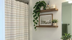 A bathroom with a striped shower curtain, a toilet with towels, a sink, a mirror, and a shelf with plants and a framed picture. - Modern Miami 2BR with Skyline Views (Miami)