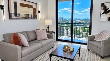A modern living room with a grey sofa, a coffee table, and a view of a cityscape through large windows.
