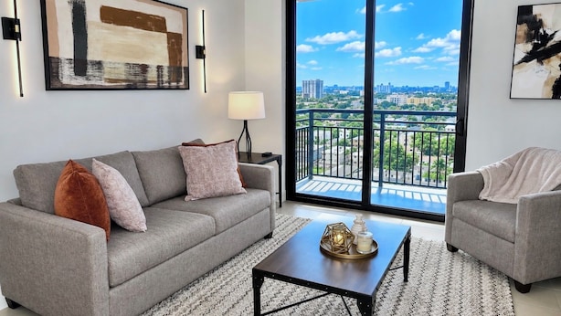 A modern living room with a grey sofa, a coffee table, and a view of a cityscape through large windows. - Modern Miami 2BR with Skyline Views (Miami)