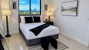 A modern bedroom with a city view, a bed with white bedding, two bedside lamps, and a framed landscape painting. - Modern Miami 2BR with Skyline Views (Miami)