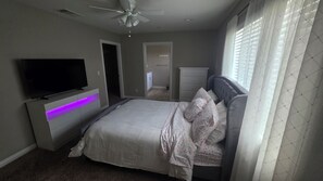 A bedroom with a bed, a TV mounted on a cabinet, a ceiling fan, and a window with blinds.