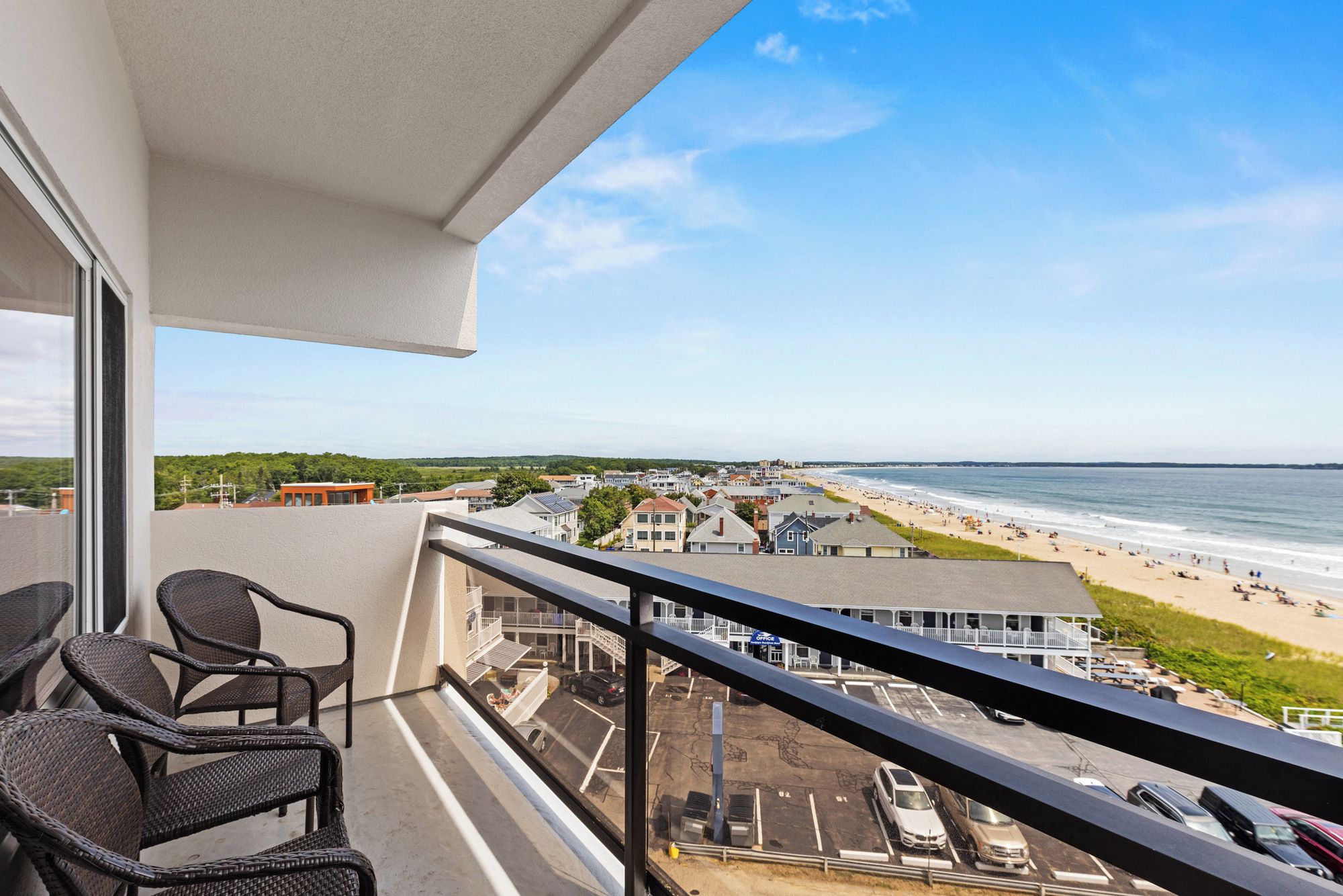 A balcony with wicker chairs, a view of a beach, and a parking lot.