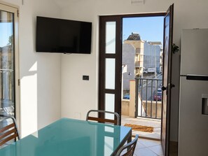 Ein Zimmer mit Glastisch, Stühlen, einem Flachbildfernseher, einer Tür zu einem Balkon und einem Kühlschrank.