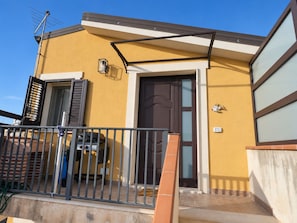 Ein Wohngebäude mit einer braunen Tür, einem kleinen Balkon mit Metallgeländer und einer sichtbaren Satellitenschüssel.