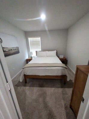 A bedroom with a bed, bedside tables, a lamp, a window with blinds, and a wall-mounted picture.