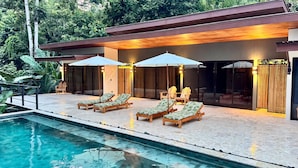 Zona de piscina exterior moderna amb gandules i ombrel·les, envoltada d'un solàrium de fusta i portes de vidre.