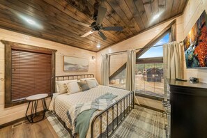 Quarto com cama, mesinha, ventilador de teto e vista para o exterior.