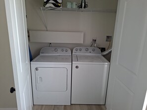 A laundry room with two white Whirlpool washers and dryers, a shelf above with laundry supplies, and a vent hose on the right. - Andy's Inn 4 Luxury Loft Apartment (Bodden Town)