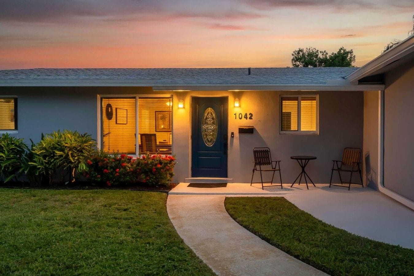 A residential house with a front door, number 1042, a small porch with two chairs, and a well-maintained lawn.