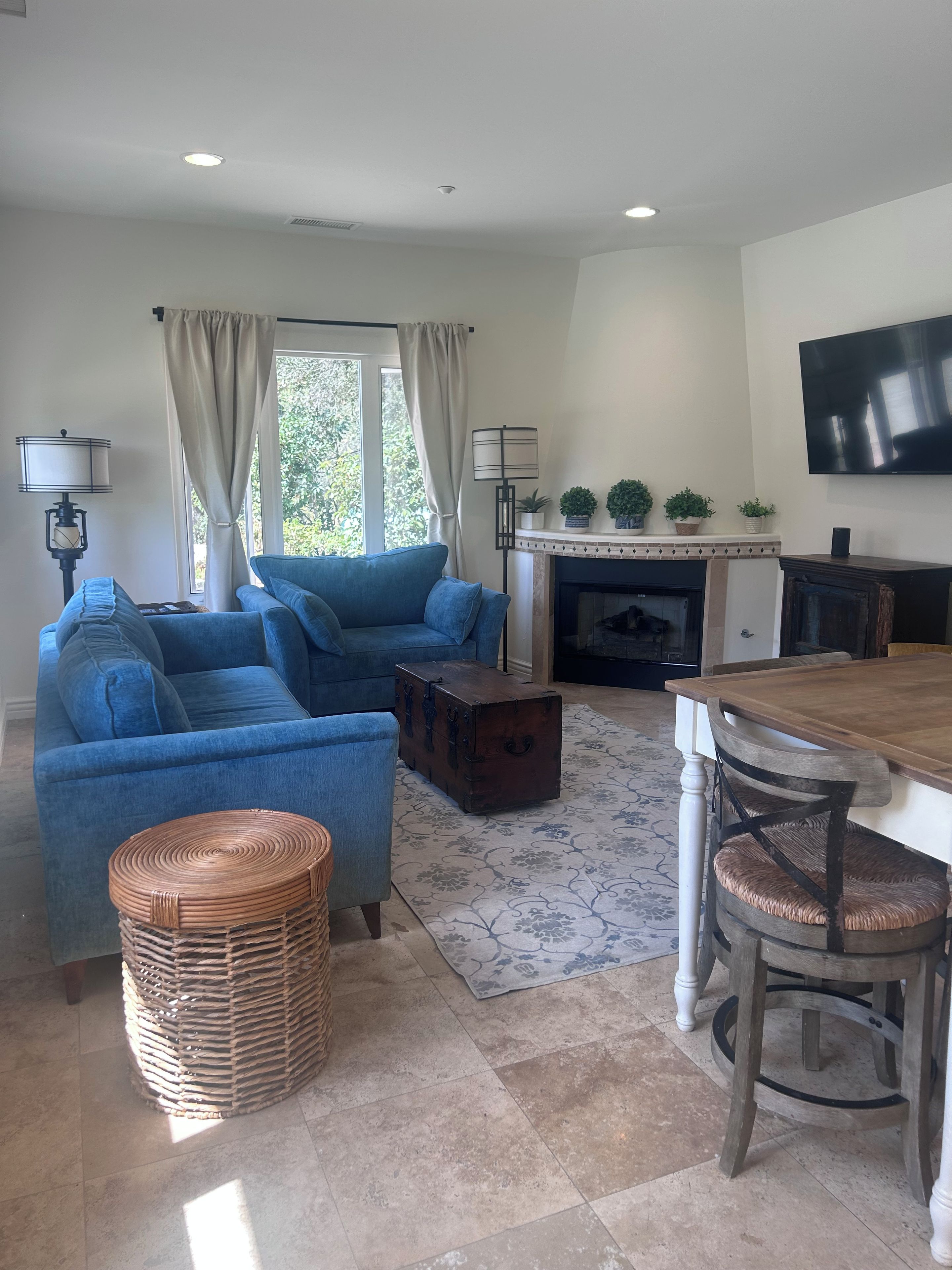 A living room with a blue sofa, a wooden chest, a dining table with chairs, a fireplace, and a television on the wall.