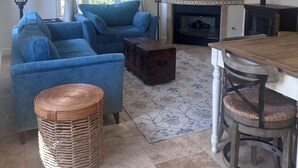 Een woonkamer met een blauwe bank, een houten kist, een eettafel met stoelen, een open haard en een televisie aan de muur.