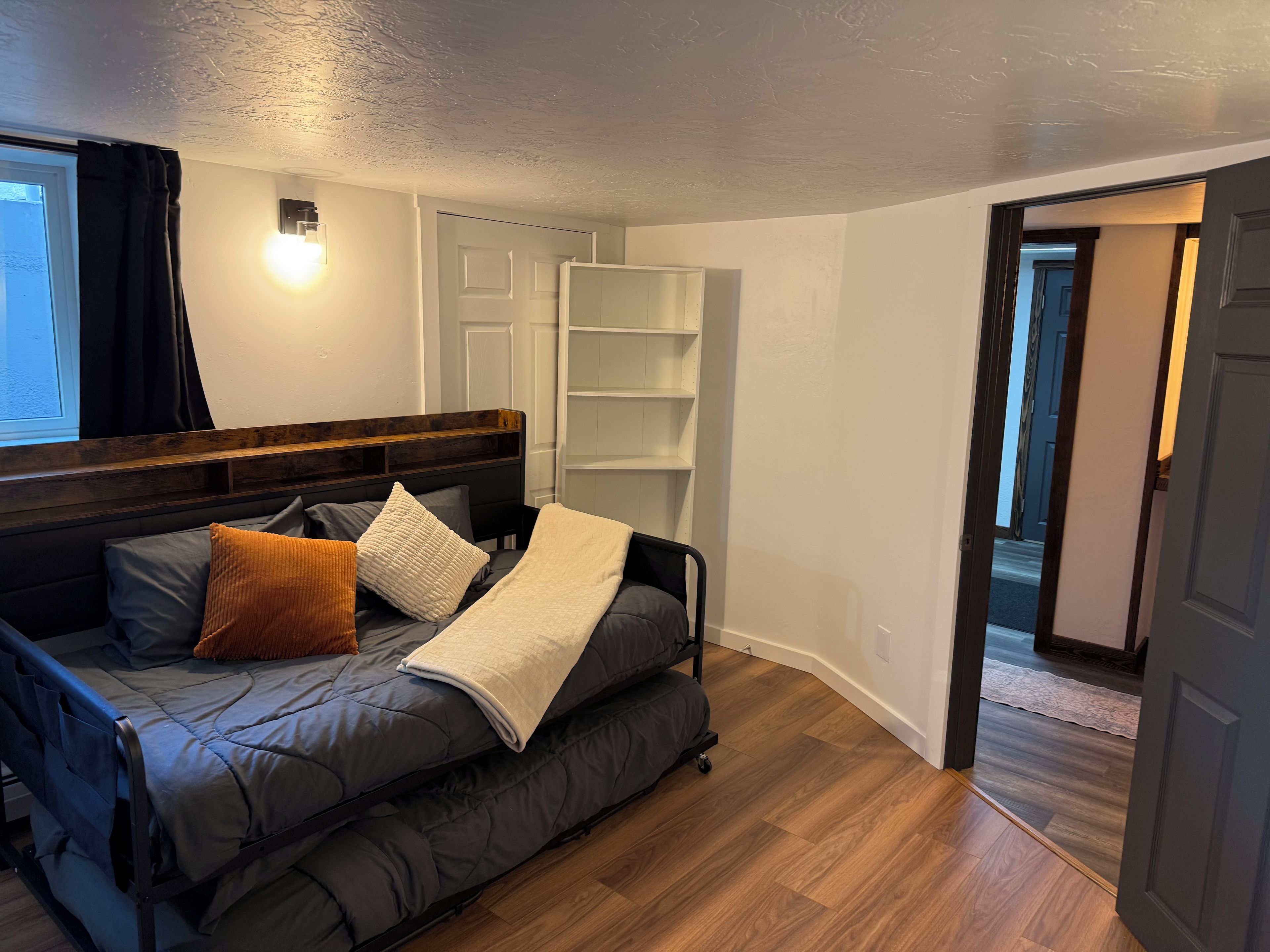 A bedroom with a bed, a nightstand, a wall-mounted shelf, and a door leading to another room.