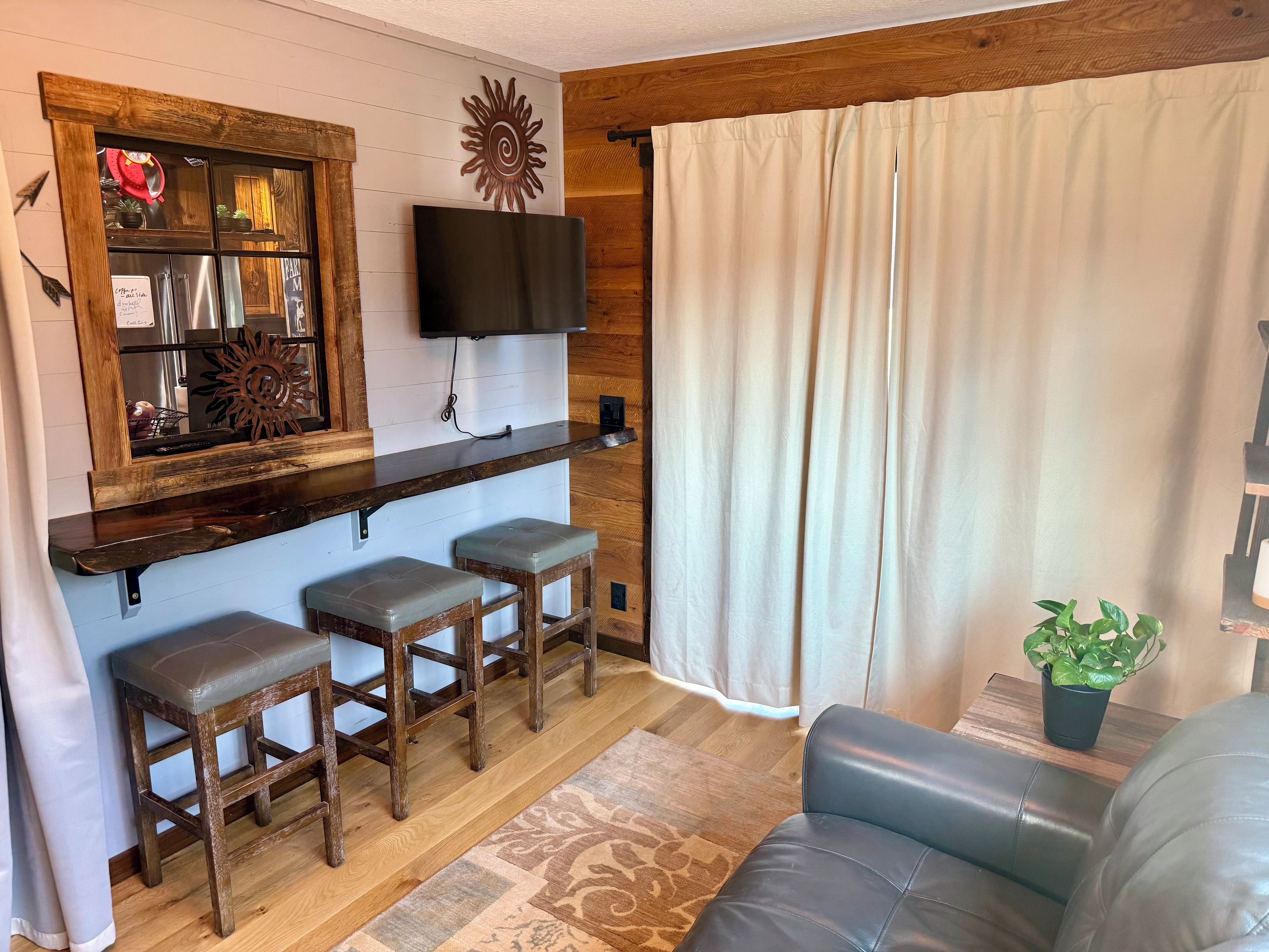 A living room with a wooden bar area, a flat-screen TV, and a large window with curtains.