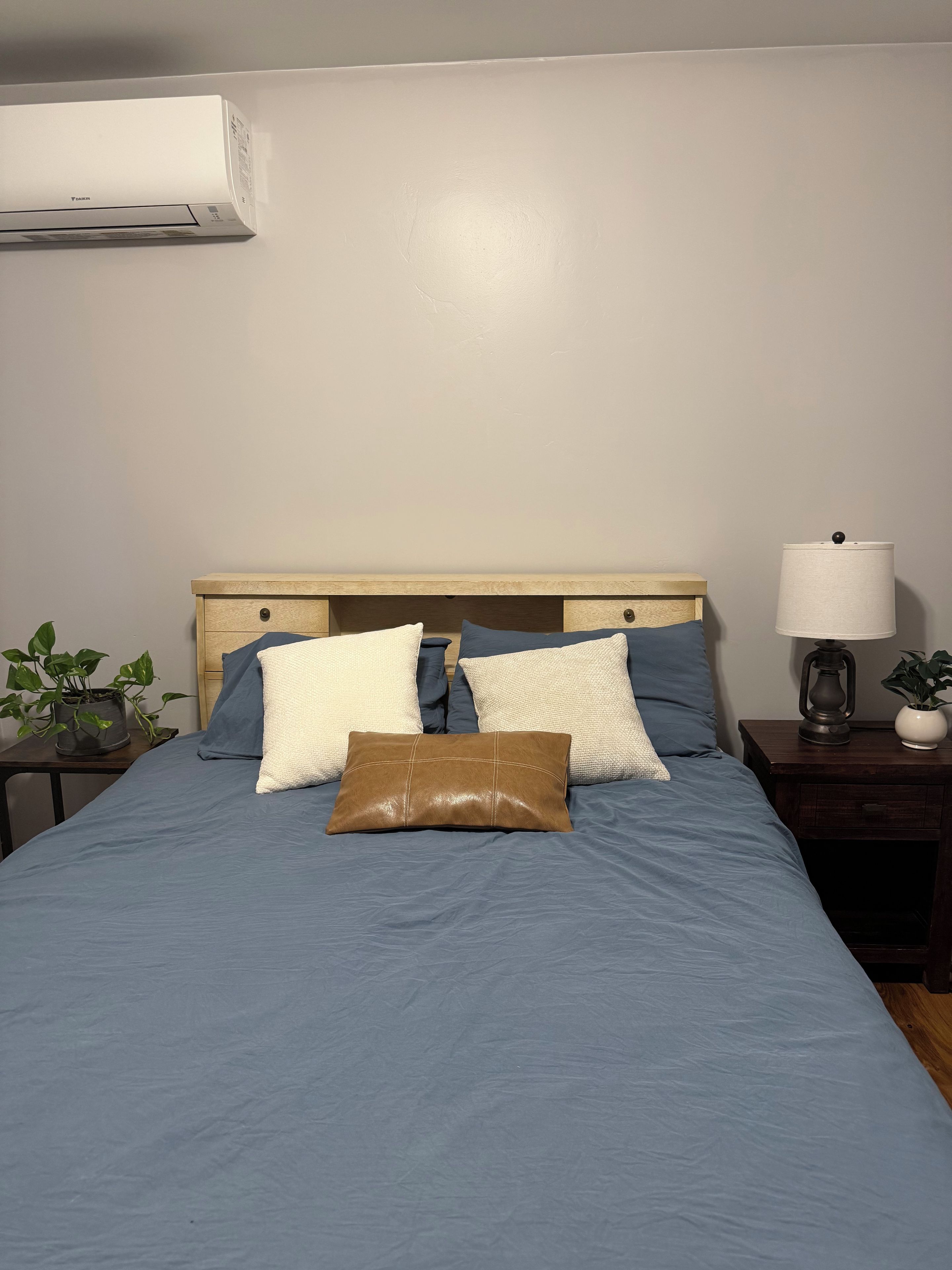 A bedroom with a bed, pillows, a nightstand, a lamp, and an air conditioning unit.