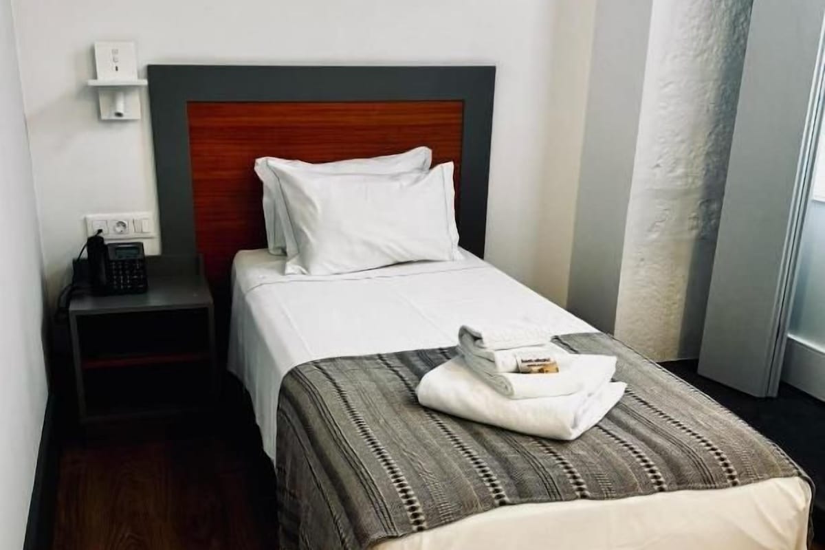 A single bed with a wooden headboard, a bedside table with a telephone, and a wall socket.