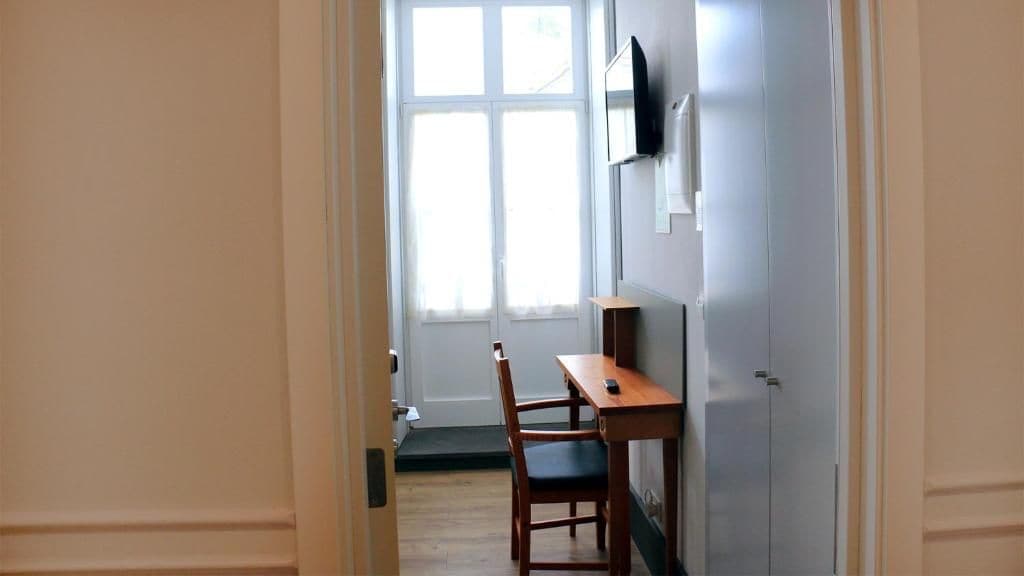 Un pasillo con un escritorio y una silla de madera, un televisor montado en la pared y una puerta que da a una habitación con ventana.