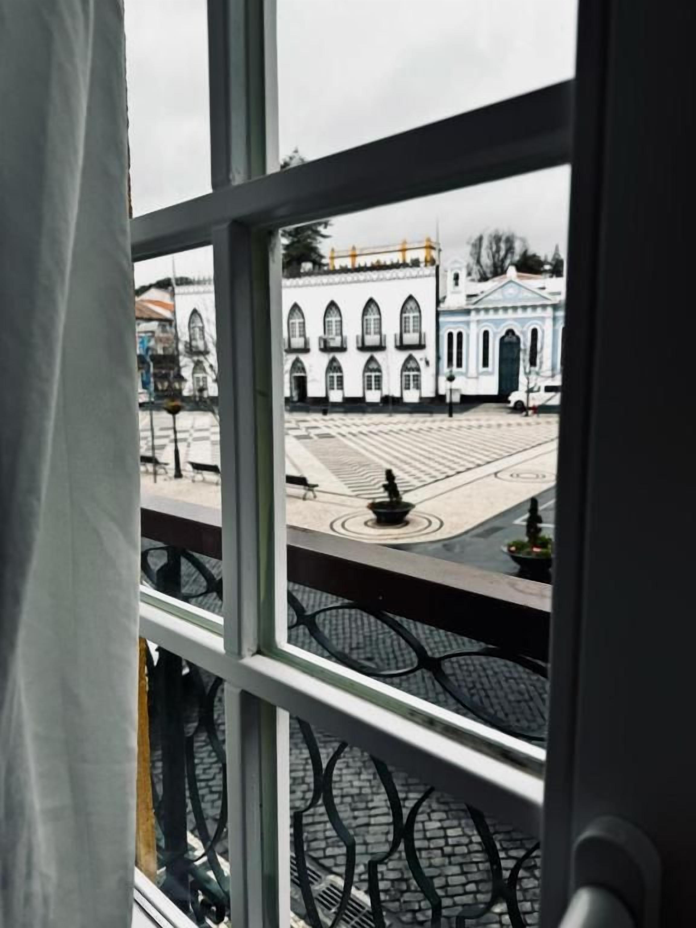 Vista pela janela, com grade de metal, para uma praça pavimentada com um edifício ao fundo.