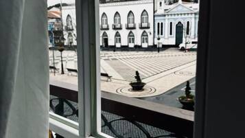Vista pela janela, com grade de metal, para uma praça pavimentada com um edifício ao fundo.