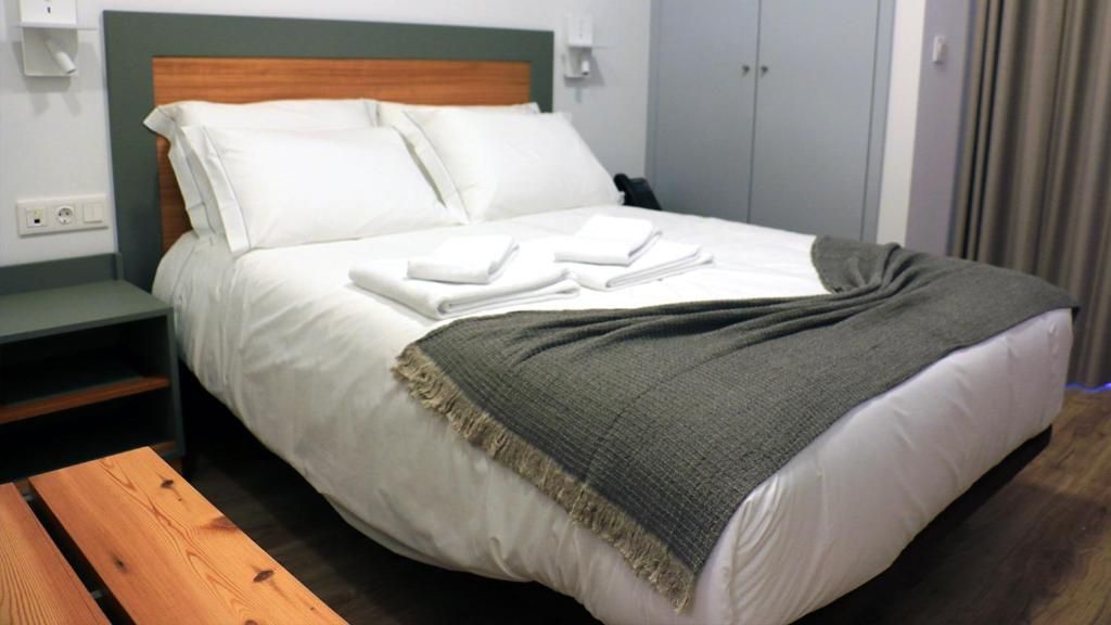 Cama bem arrumada com lençóis brancos e manta cinza, mesa de cabeceira com abajur e um banco de madeira.