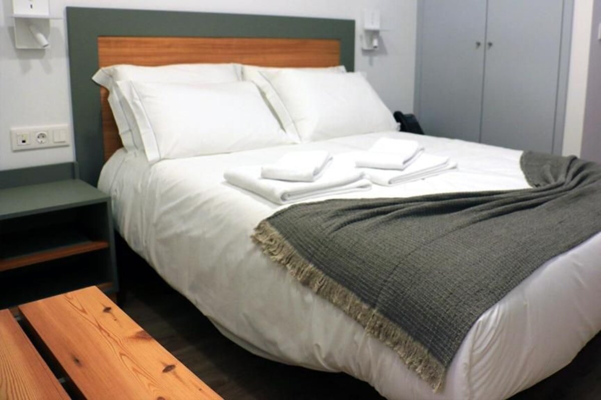A neatly made bed with white linens and a grey throw blanket, a bedside table with a lamp, and a wooden bench.