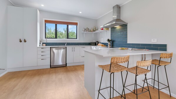 A modern kitchen with white cabinetry, a built-in dishwasher, a large island with bar stools, and a window with a view of greenery.