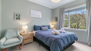 Un dormitorio con cama, mesitas de noche, una silla y ventana con vista a árboles.