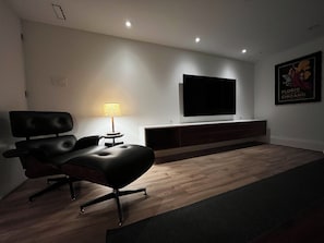 A modern living room with a black leather sofa, a round table with a lamp, a flat-screen TV mounted on the wall, and a wooden floor. - Nº577 – Private Boutique Garden Suite in the Annex, Steps to Bloor & Subway. (Toronto)