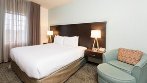 Habitación de hotel con una cama grande, dos lámparas de noche, una mesita de noche con teléfono y un sillón pequeño con cojín estampado.