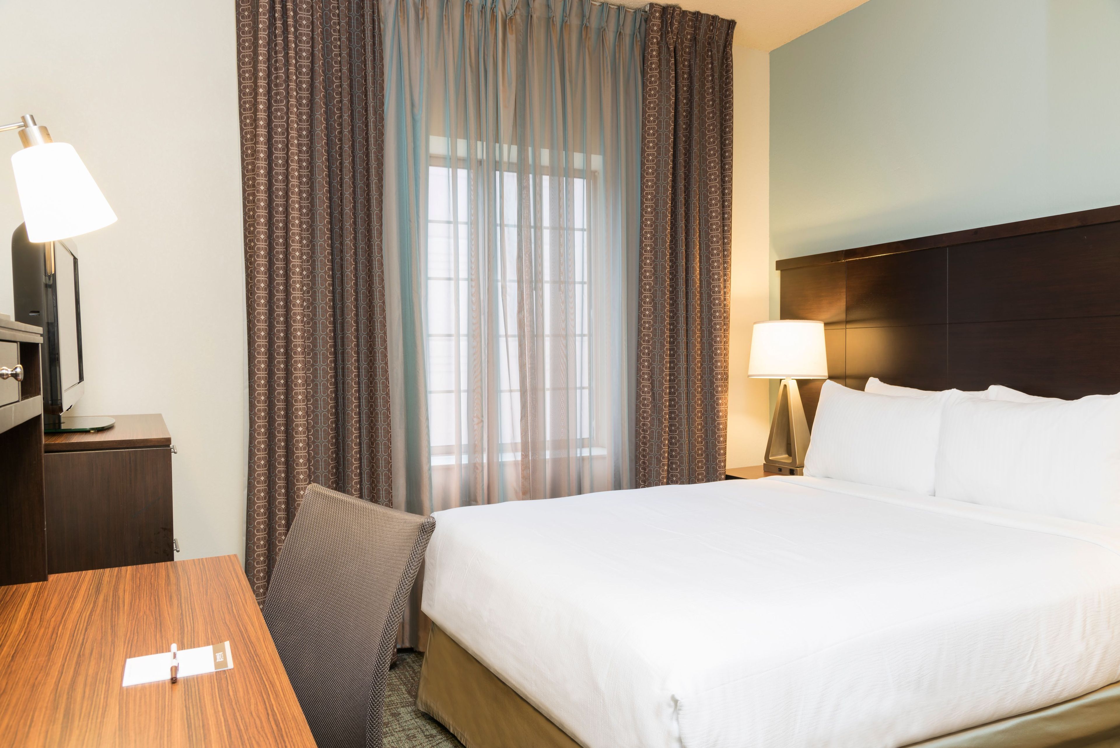 A hotel room with a bed, a desk, a chair, a lamp, and curtains.