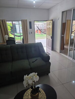 En stue med sofa, et sofabord med orkideer og udsigt gennem en åben dør til en patio.