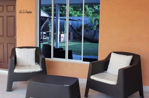 Un patio con mobiliario de mimbre, incluyendo dos sillas y una mesa, y un ventanal con vista a un jardín trasero.