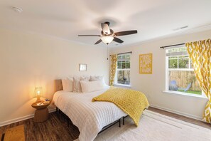 Quarto com cama, mesa de cabeceira, abajur, ventilador de teto, janelas e vista para o exterior.