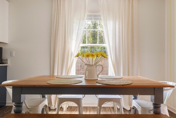 Uma mesa de jantar com um jarro branco e flores amarelas, rodeada por cadeiras brancas.