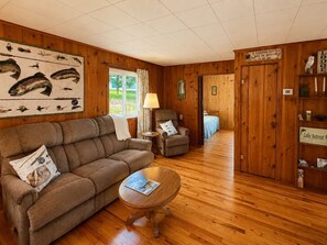 Interior acogedor de una cabaña con piso de madera, un sofá, una silla, una mesita y una cama al fondo.