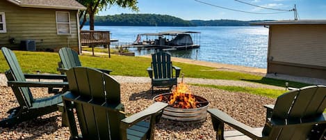 Un área de estar al aire libre junto al lago con sillas Adirondack, un brasero y vistas a un muelle y barcos.