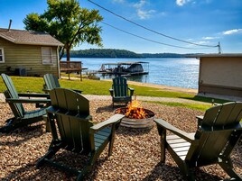 Un área de estar al aire libre junto al lago con sillas Adirondack, un brasero y vistas a un muelle y barcos.