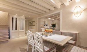 A dining area with a white table, chairs, and a bench. There is a wall-mounted light fixture and a kitchen in the background. - BEAUTIFUL 3-BEDROOM DORSODURO APARTMENT WITH PRIVATE GARDEN (Venezia)