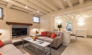 A living room with a fireplace, a sofa, a coffee table with magazines, and a kitchen area visible through an open doorway. - BEAUTIFUL 3-BEDROOM DORSODURO APARTMENT WITH PRIVATE GARDEN (Venezia)