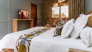 Une chambre à coucher avec un lit, des oreillers, un téléviseur posé sur un meuble, une fenêtre avec des rideaux en bambou et un luminaire suspendu.