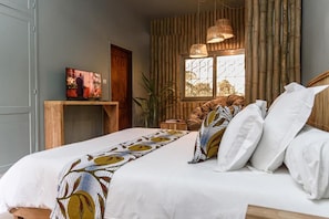 Een slaapkamer met een bed, kussens, een televisie op een standaard, een raam met bamboesoorten gordijnen en een hanglamp.