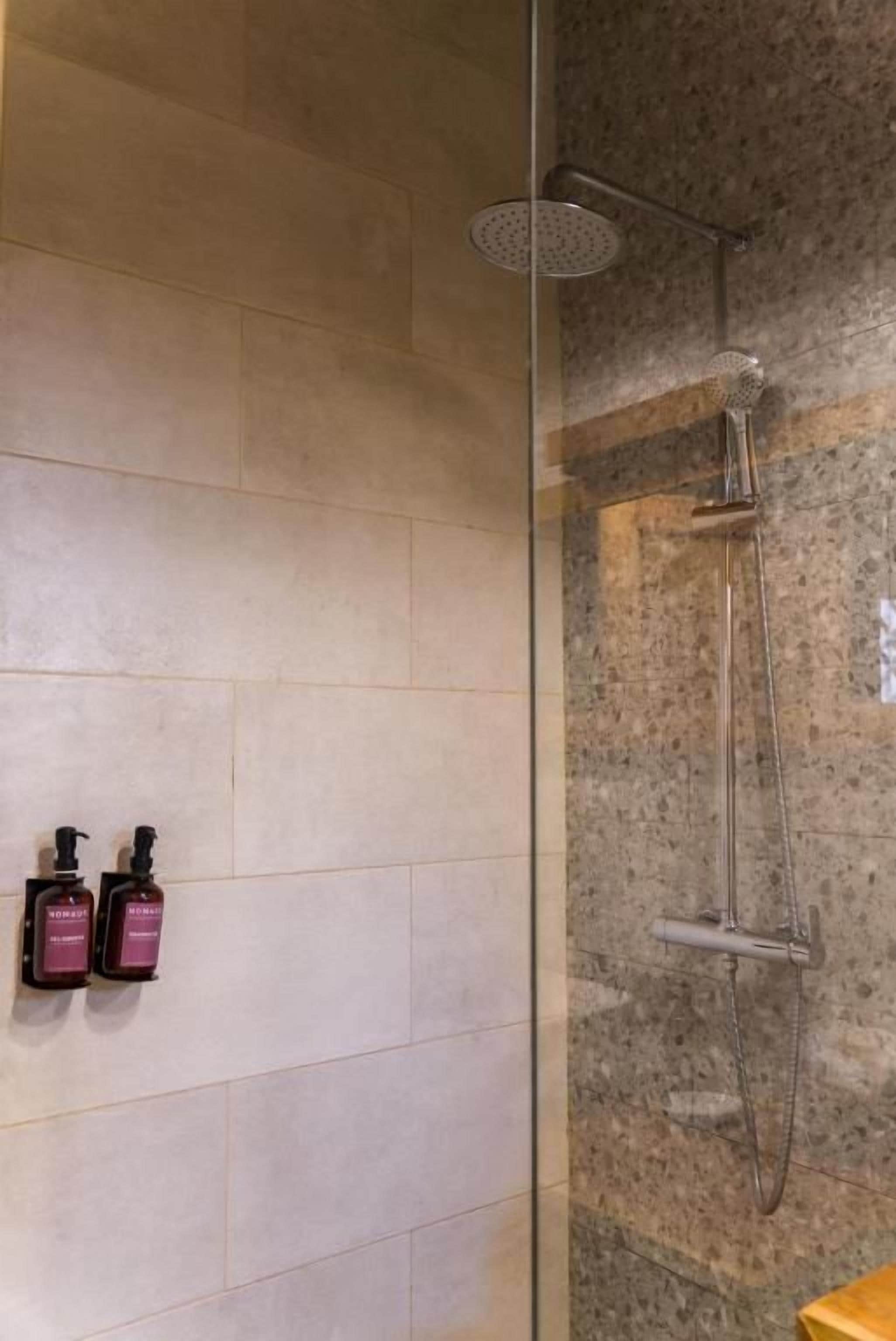 A shower area with a glass partition, a handheld showerhead, and two bottles of shampoo.