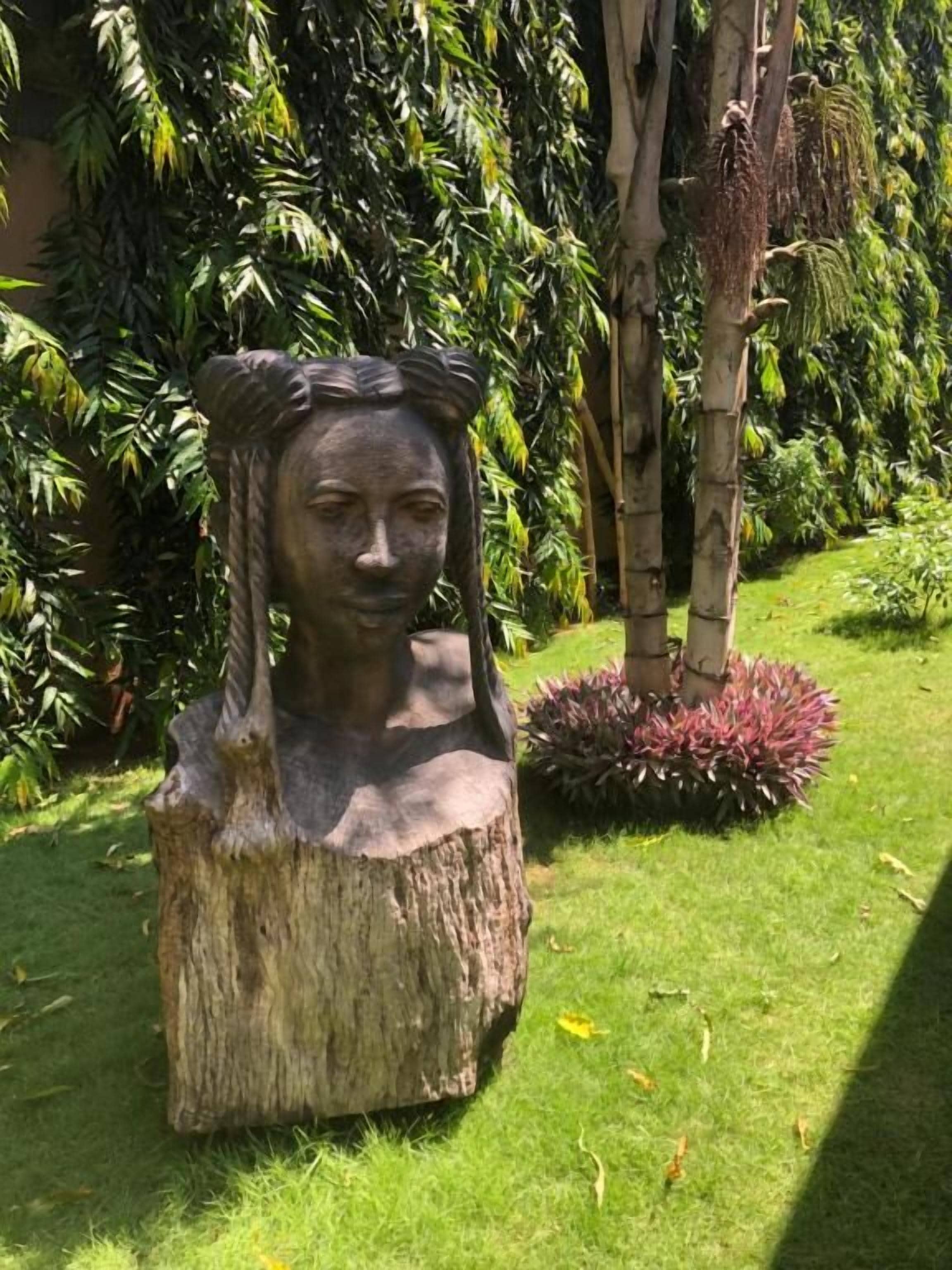 Escultura de una persona sobre un tocón en un jardín con follaje exuberante y un pequeño arbusto.