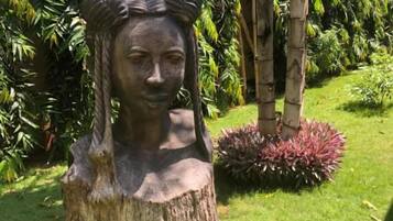 Escultura de una persona sobre un tocón en un jardín con follaje exuberante y un pequeño arbusto.