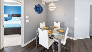 Una habitación de hotel moderna con una mesa de comedor de vidrio, sillas blancas, una pared azul de acento y una lámpara en el techo.
