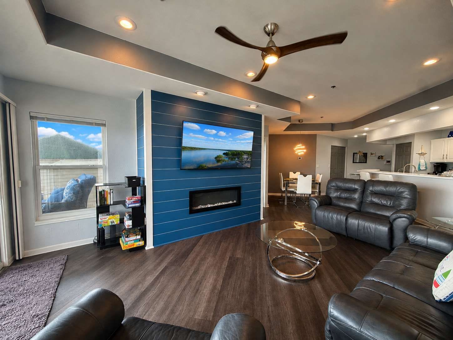 A modern living room with a flat-screen TV, leather sofas, a glass coffee table, and a ceiling fan.