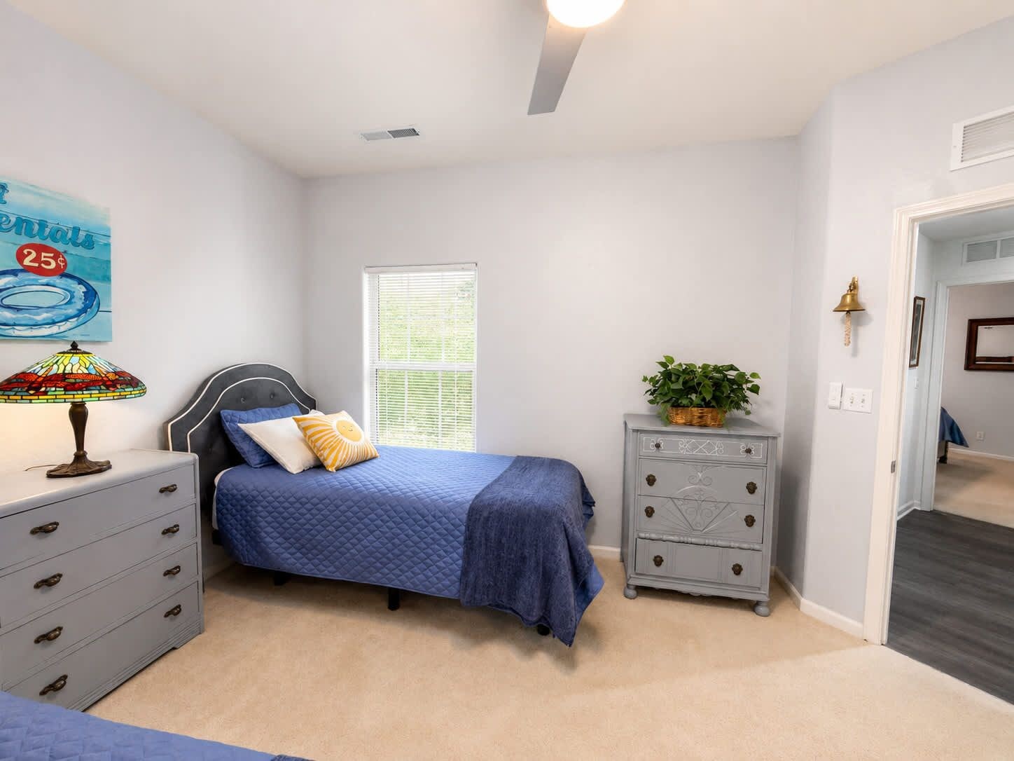 Un dormitorio con cama, cómoda, mesita de noche, lámpara, ventana y puerta que da a otra habitación.
