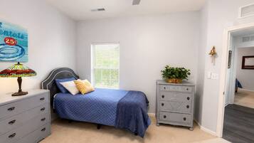 Un dormitorio con cama, cómoda, mesita de noche, lámpara, ventana y puerta que da a otra habitación.