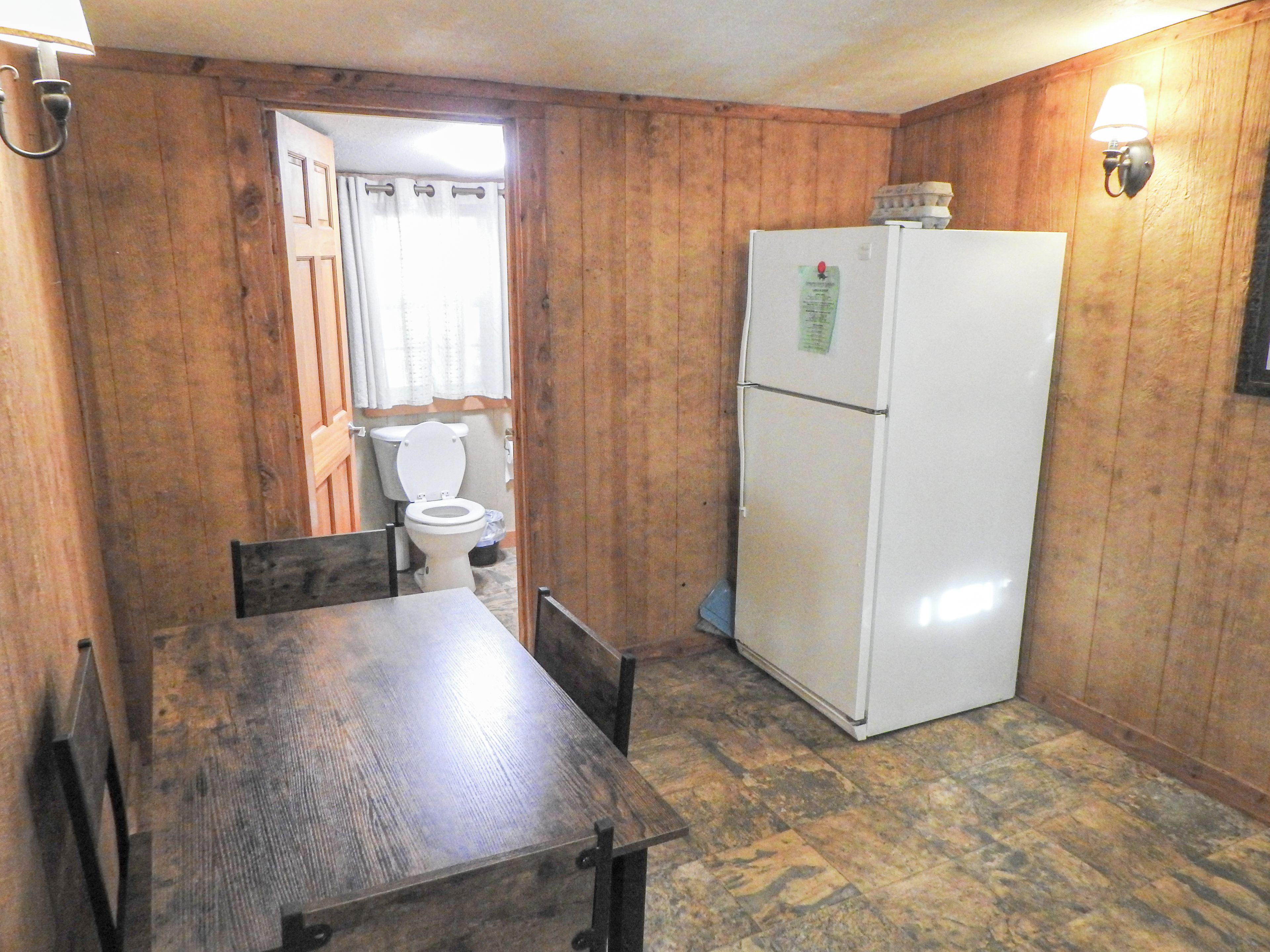 A compact kitchen with wooden walls, a white refrigerator, a wooden dining table, and a basic toilet.