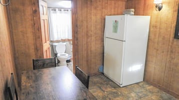 A compact kitchen with wooden walls, a white refrigerator, a wooden dining table, and a basic toilet.